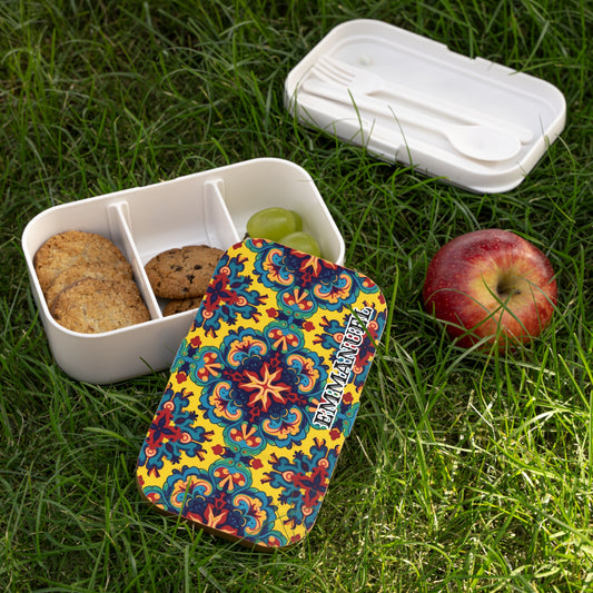 A colorful African-inspired bento lunch box with a vibrant floral pattern sits open on green grass. Inside, there are two cookies, one chocolate chip cookie, and two green grapes. A red apple and white cutlery in a case are beside the lunchbox.