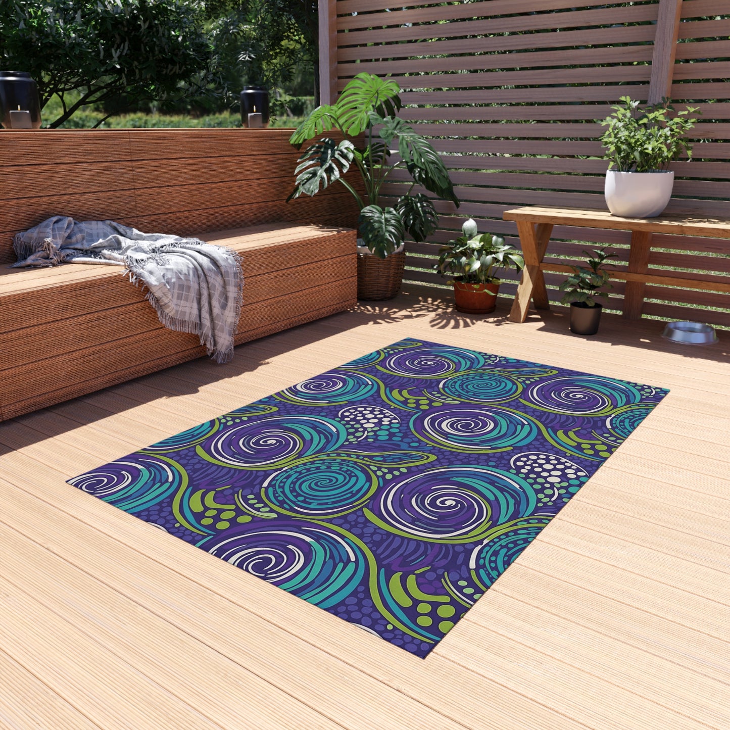 A modern patio with wooden decking features a colorful, weather-resistant rug in swirling blue, green, and purple patterns. There’s a wicker bench with a gray blanket, a wooden bench, potted plants, and horizontal slat fencing in the background.
