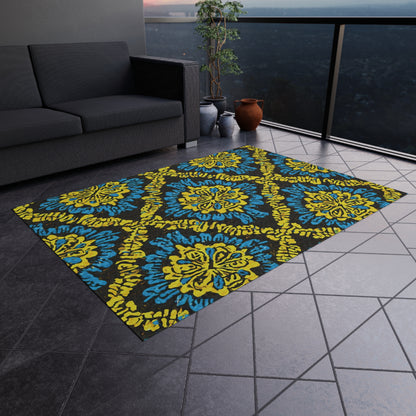 A modern balcony features a gray sectional sofa, an African-inspired outdoor rug with blue-and-yellow ornate patterns, two clay pots with leafy plants, and dark tiled flooring. A glass railing offers a cityscape view at dusk.