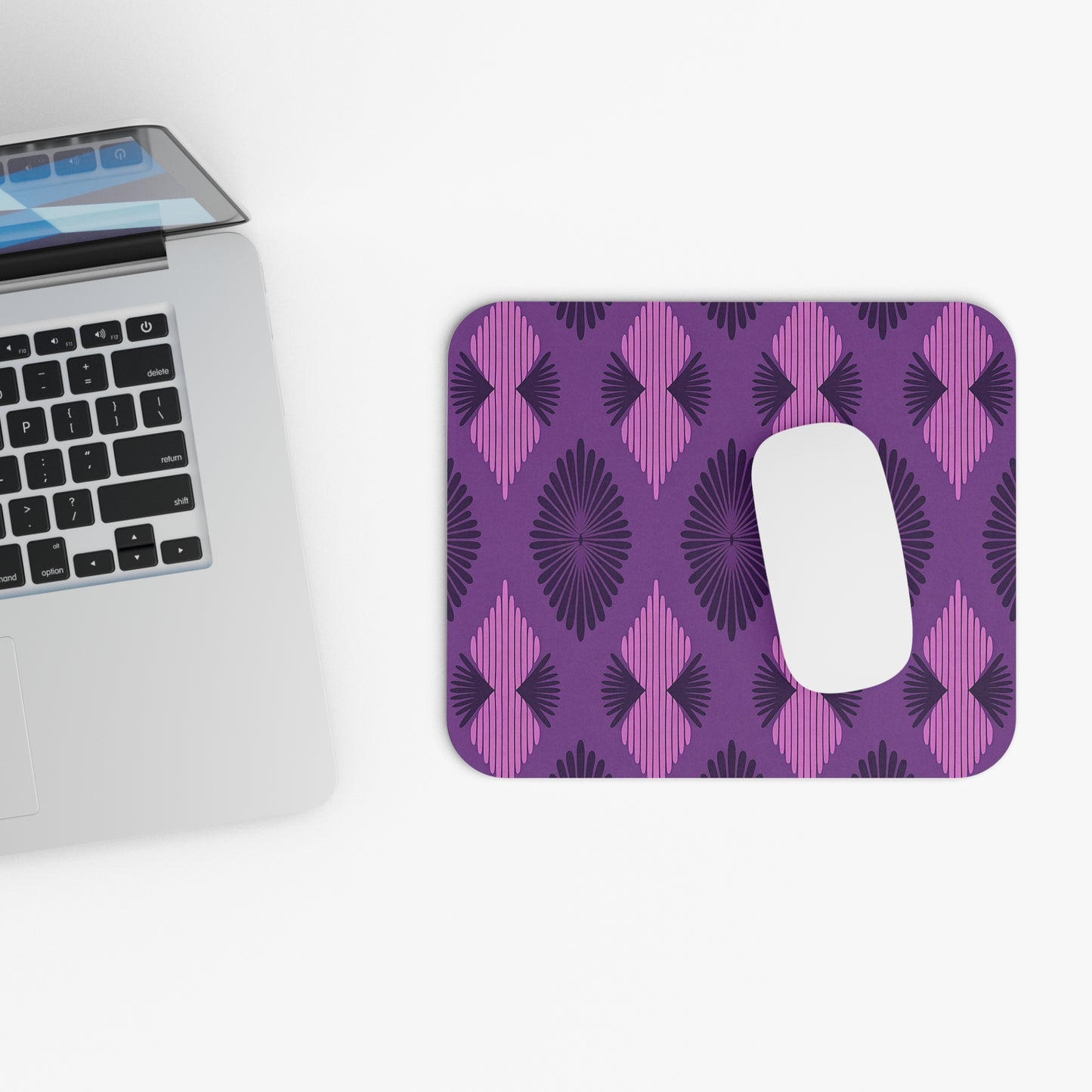 A silver laptop sits on a white surface next to a vibrant desk accessory—a rectangular mouse pad with modern purple and magenta geometric patterns, radiating black lines, and a non-slip rubber base—plus a white mouse on top.