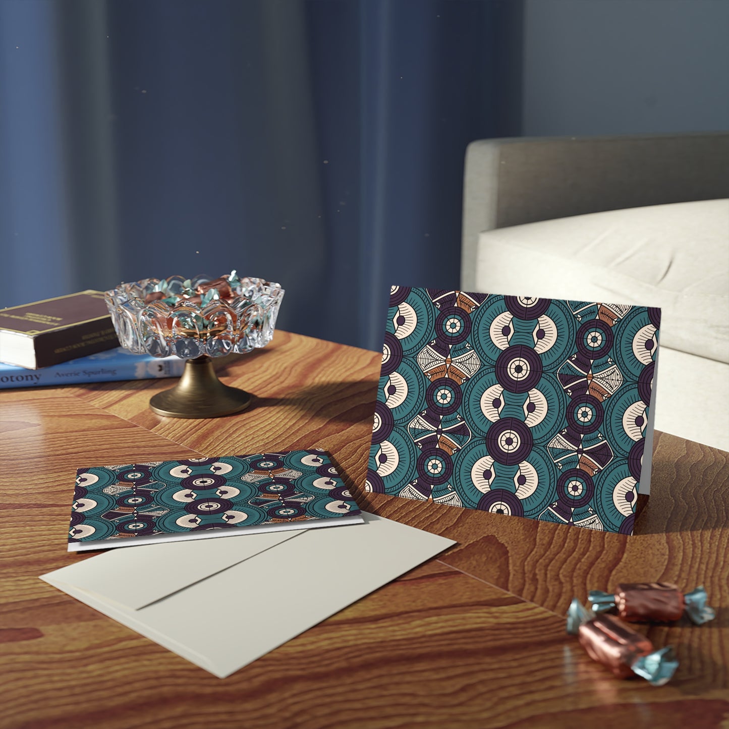 A patterned, African-inspired greeting card and matching envelope sit on a wooden table beside two blank cards. A glass dish of chocolates and books rest nearby. Sunlight streams in, casting shadows, with a gray sofa and blue curtain in the background.