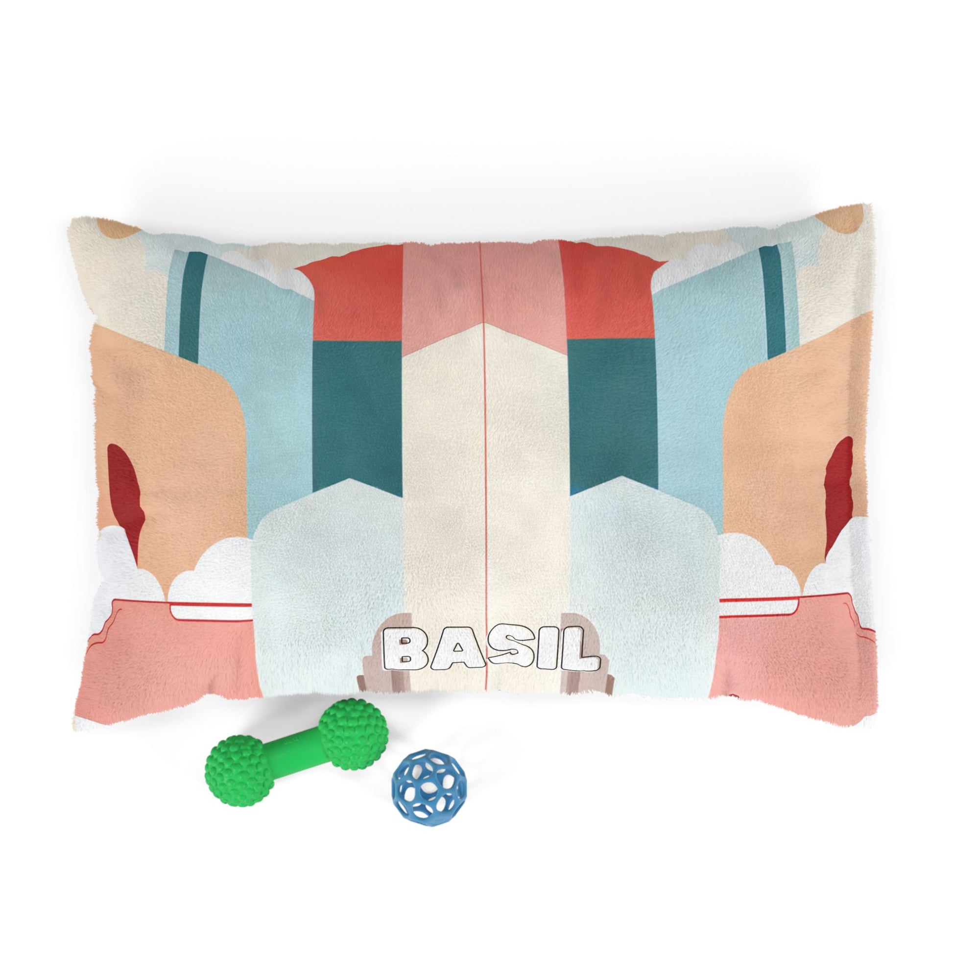 A rectangular pet bed with an African-inspired, abstract geometric design in teal, coral, beige, and white features the word “BASIL” in bold at the front. Two dog toys—a green dumbbell and a blue ball—lie before it on a white background.