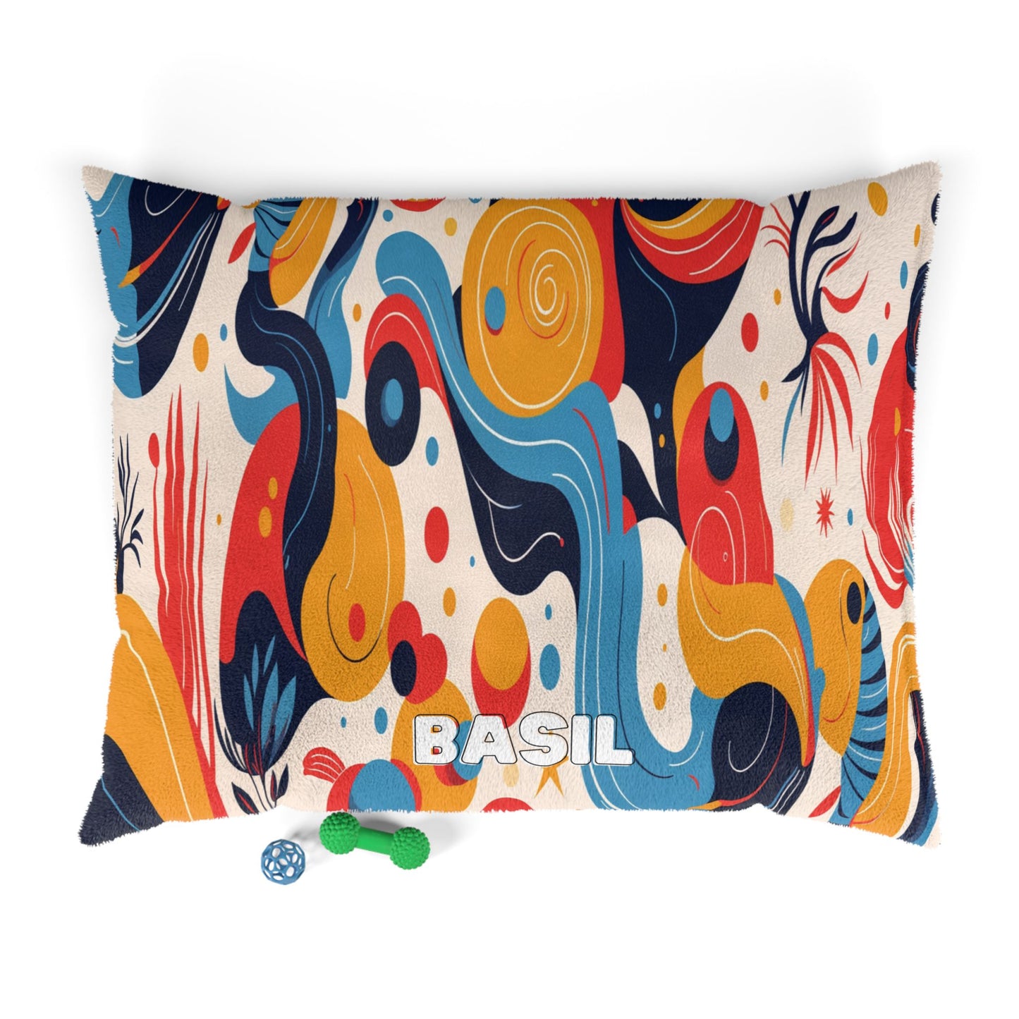 A rectangular pillow with “BASIL” embroidered in white block letters features a playful, African-inspired abstract pattern in blue, red, orange, and yellow. On the floor below are a green bone-shaped toy and a blue ball with paw prints.