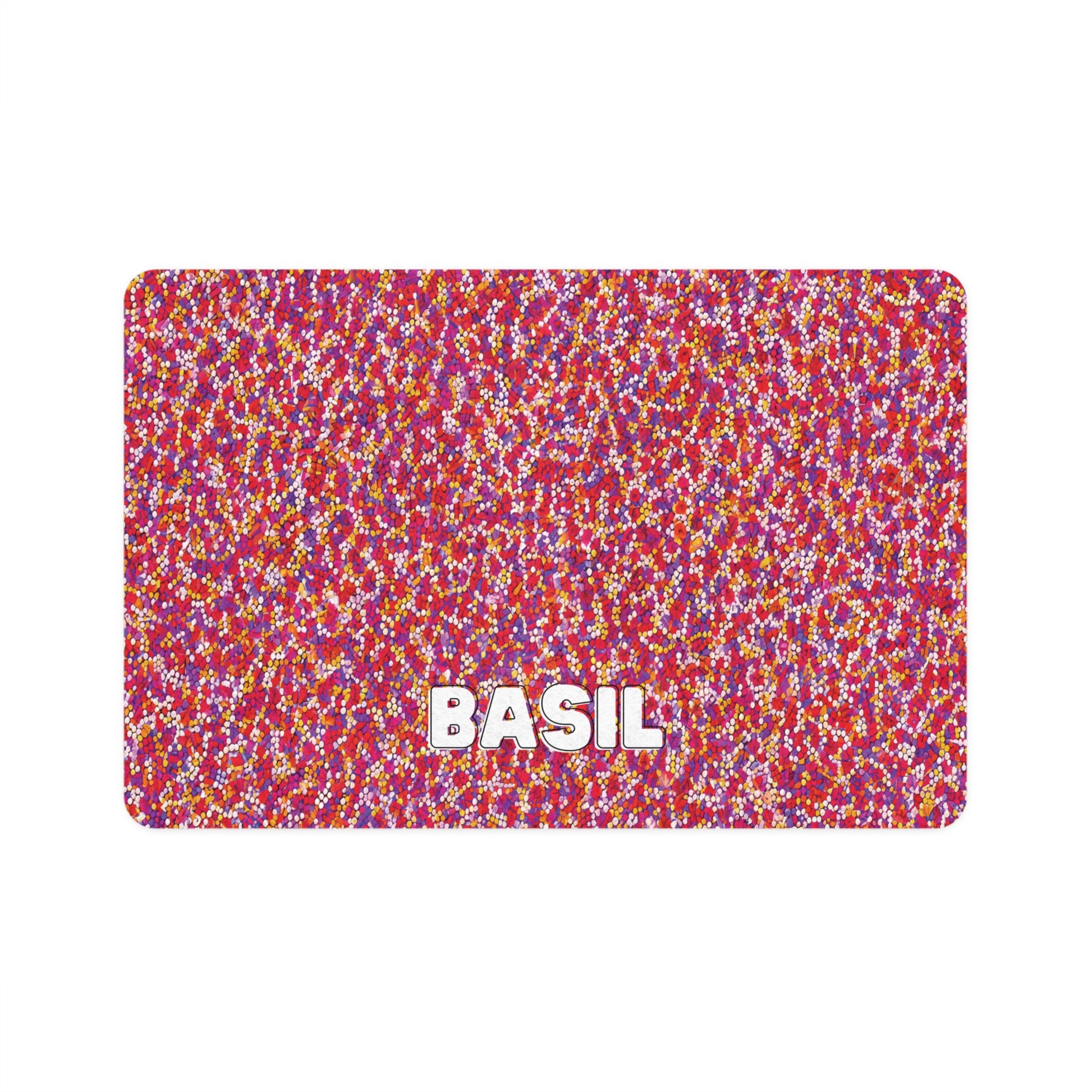 A rectangular mat featuring an African-inspired dense pattern of small red, pink, yellow, and white dots. “BASIL” appears in large, bold white letters with a gray shadow near the bottom center, set against a plain white background.