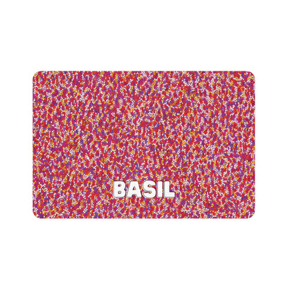 A rectangular mat featuring an African-inspired dense pattern of small red, pink, yellow, and white dots. “BASIL” appears in large, bold white letters with a gray shadow near the bottom center, set against a plain white background.