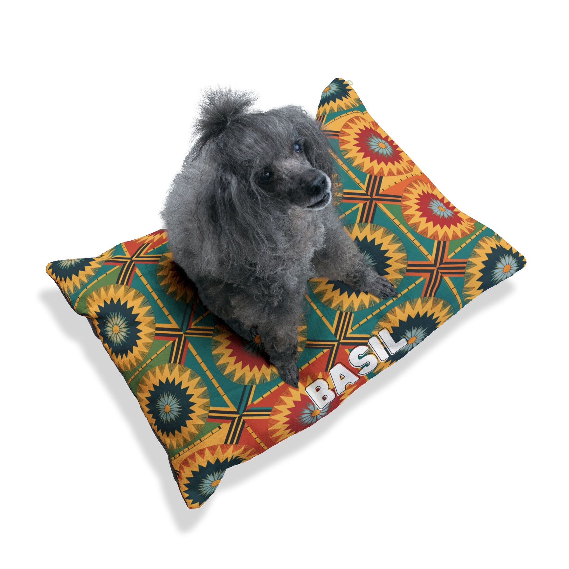 A small gray poodle sits on a colorful cushion with a geometric starburst pattern in green, orange, red, and yellow. The personalized pet bed has the name BASIL in bold white letters. The background is plain white.