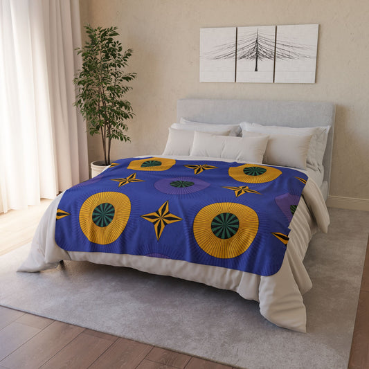 A neatly made bed with a blue sherpa fleece blanket featuring large circular yellow and green patterns sits in a bright, modern bedroom. A potted tree is by the window with white curtains, and minimalist black-and-white artwork hangs above the bed.