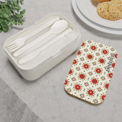 A white personalized bento box with a floral-patterned lid and the word EXEMPLARY sits on a light gray surface. Next to it, white plastic fork, spoon, and knife are neatly arranged with cookies and green leaves nearby.