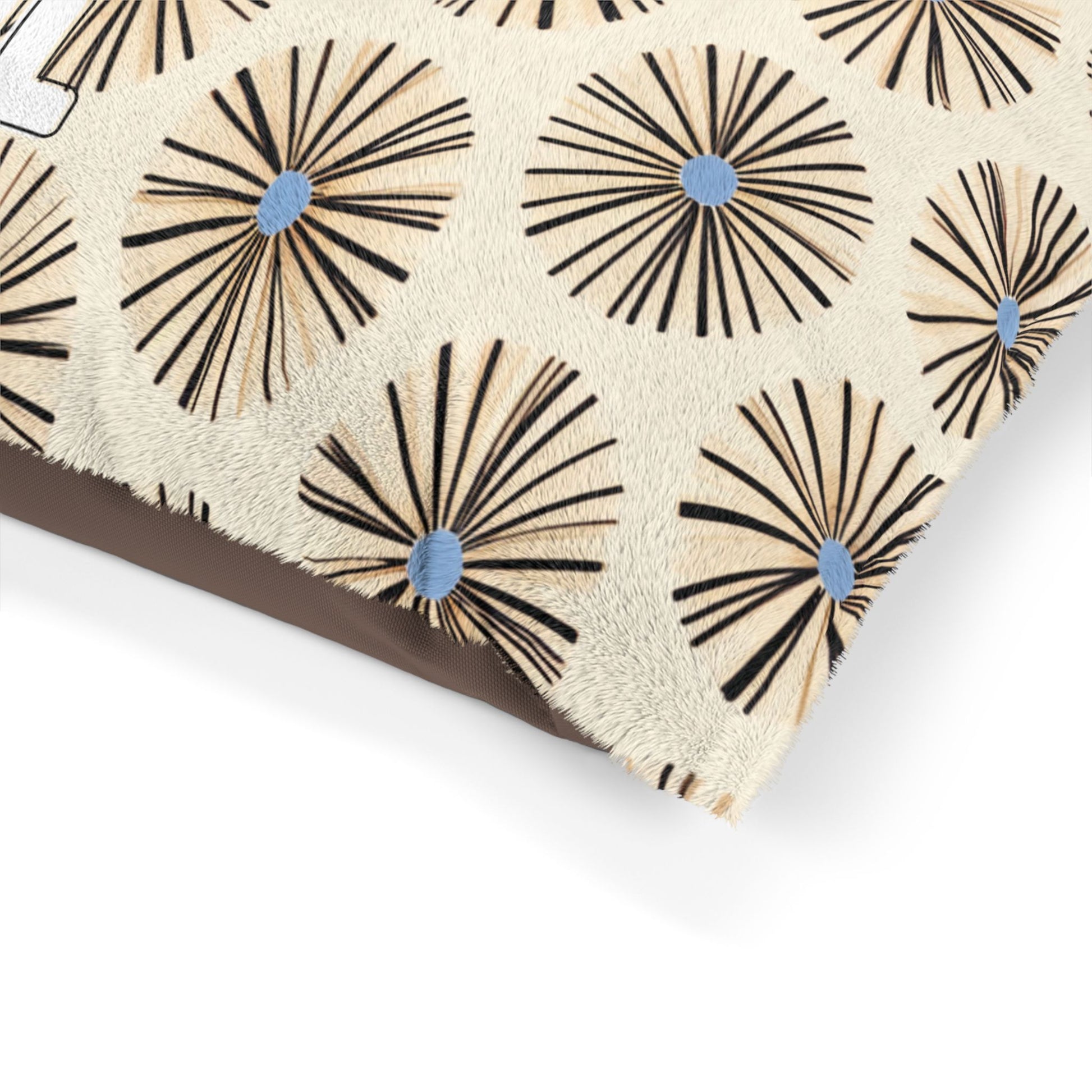 A close-up of a beige pillow with a textured surface, featuring a repeating geometric pattern of circular bursts. Each burst has black and tan radiating lines with a blue dot at the center, evoking an African-inspired, mid-century modern design.