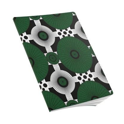 A closed African-inspired journal with a geometric cover design featuring large green circular shapes with radiating lines, black concentric circles, and black dots on a white background. The notebook is angled and pages for daily journaling are slightly visible.