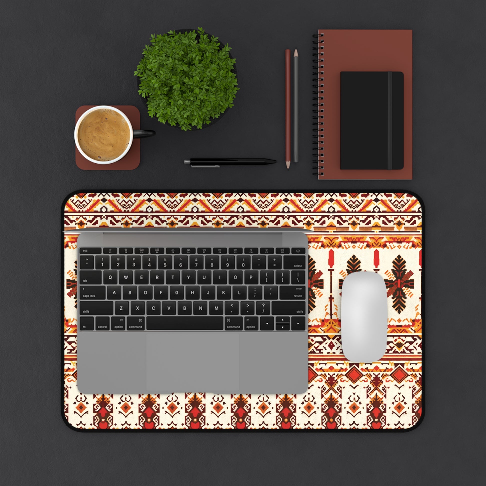 A top-down view of a workspace featuring a laptop on an ergonomic Neoprene desk mat with red and brown geometric designs, a white mouse, coffee cup, small green potted plant, two pens, and two brown notebooks arranged neatly on a dark surface.