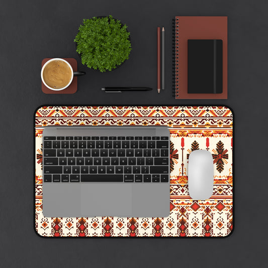 A top-down view of a workspace featuring a laptop on an ergonomic Neoprene desk mat with red and brown geometric designs, a white mouse, coffee cup, small green potted plant, two pens, and two brown notebooks arranged neatly on a dark surface.