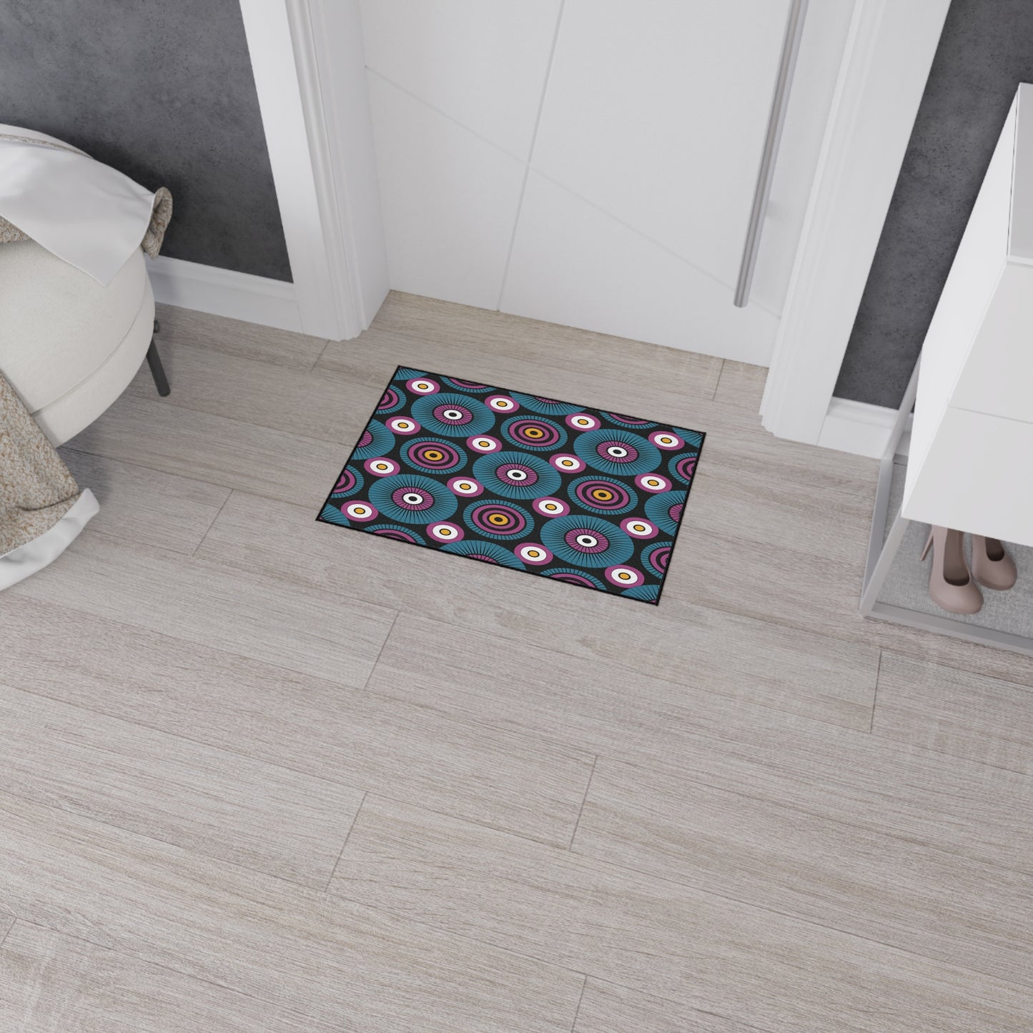 A small rectangular doormat with a colorful, African-inspired circular pattern and non-slip rubber backing lies on light wood flooring in front of a white door, near a white chair, shoe rack, and a section of black accent wall.