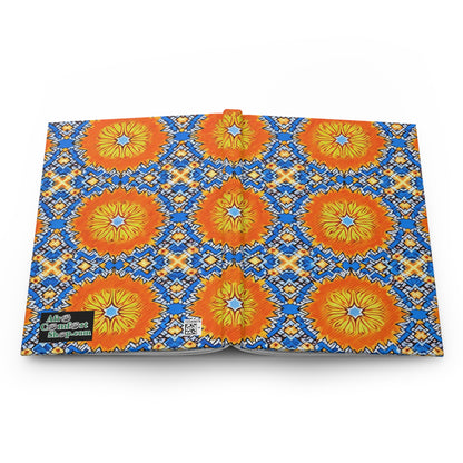Open book displaying a vibrant pattern of orange and yellow lion faces with blue and yellow sunburst designs on a white background. The back cover features a green “Afro Comix Print” logo and QR code, perfect for an African-inspired journal.