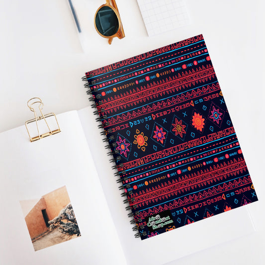 A spiral notebook with a dark cover featuring bright, culturally inspired geometric and floral patterns lies on a white desk. Nearby are an open book displaying a photo, gold binder clip, sunglasses, and a small grid notepad.