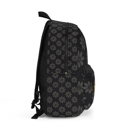 A black boho chic rucksack viewed from the side, featuring a repeating pattern of gray daisy-like flowers. The bag has a front pocket, mesh side pocket, zippers on top, and adjustable shoulder straps visible on the left. Plain white background.