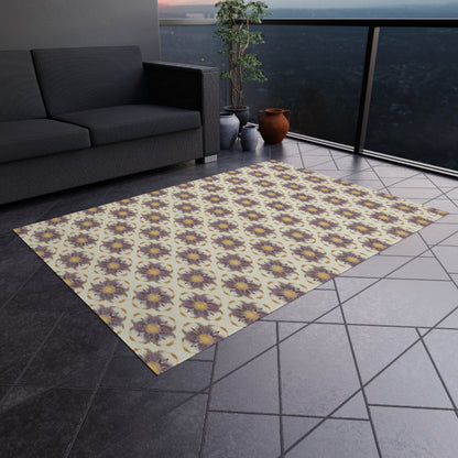 An African-inspired patterned rug with yellow and purple flower motifs lies on a dark-tiled balcony floor. A dark gray sofa, potted plant, and clay pots are nearby, with a hazy cityscape visible beyond the glass balcony railing.