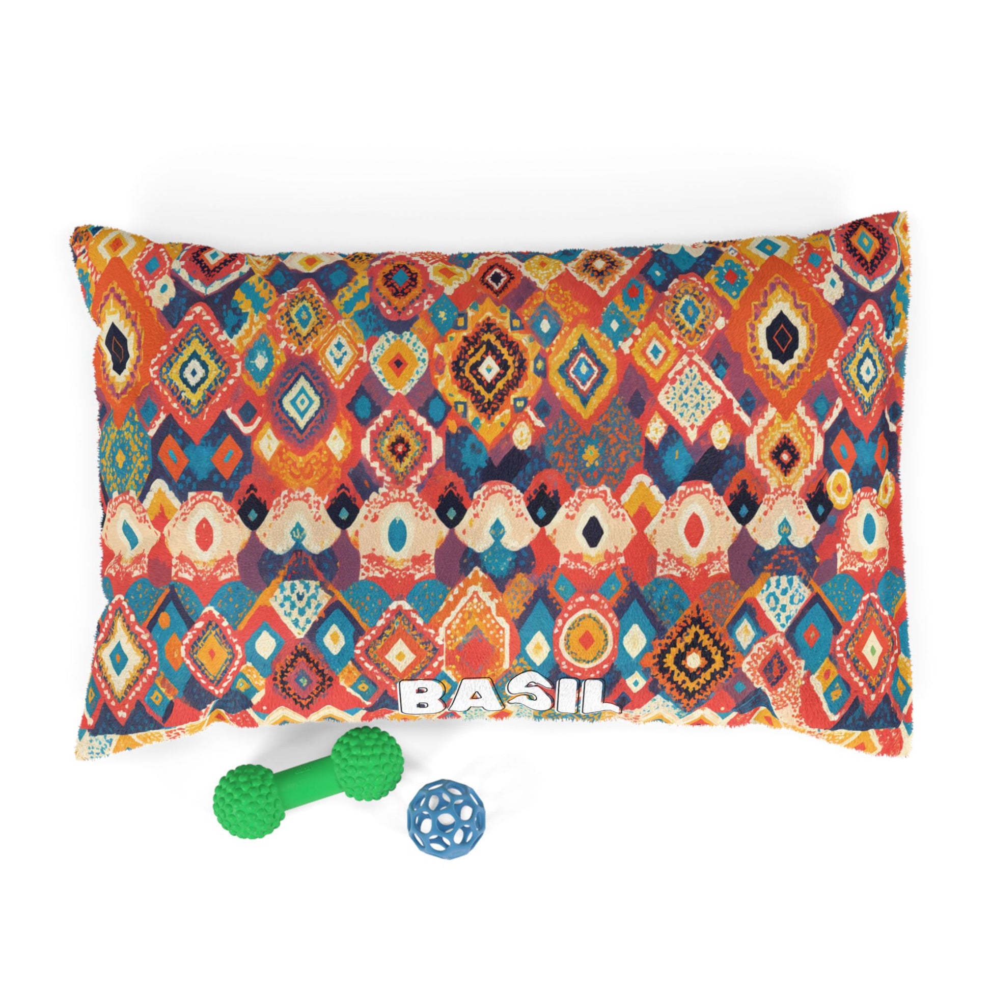 A rectangular dog bed with an African-inspired, colorful geometric diamond pattern in red, orange, blue, and cream. The name BASIL is printed in white letters at the bottom. Two dog toys—a green double-ball and a blue lattice ball—sit below the bed.