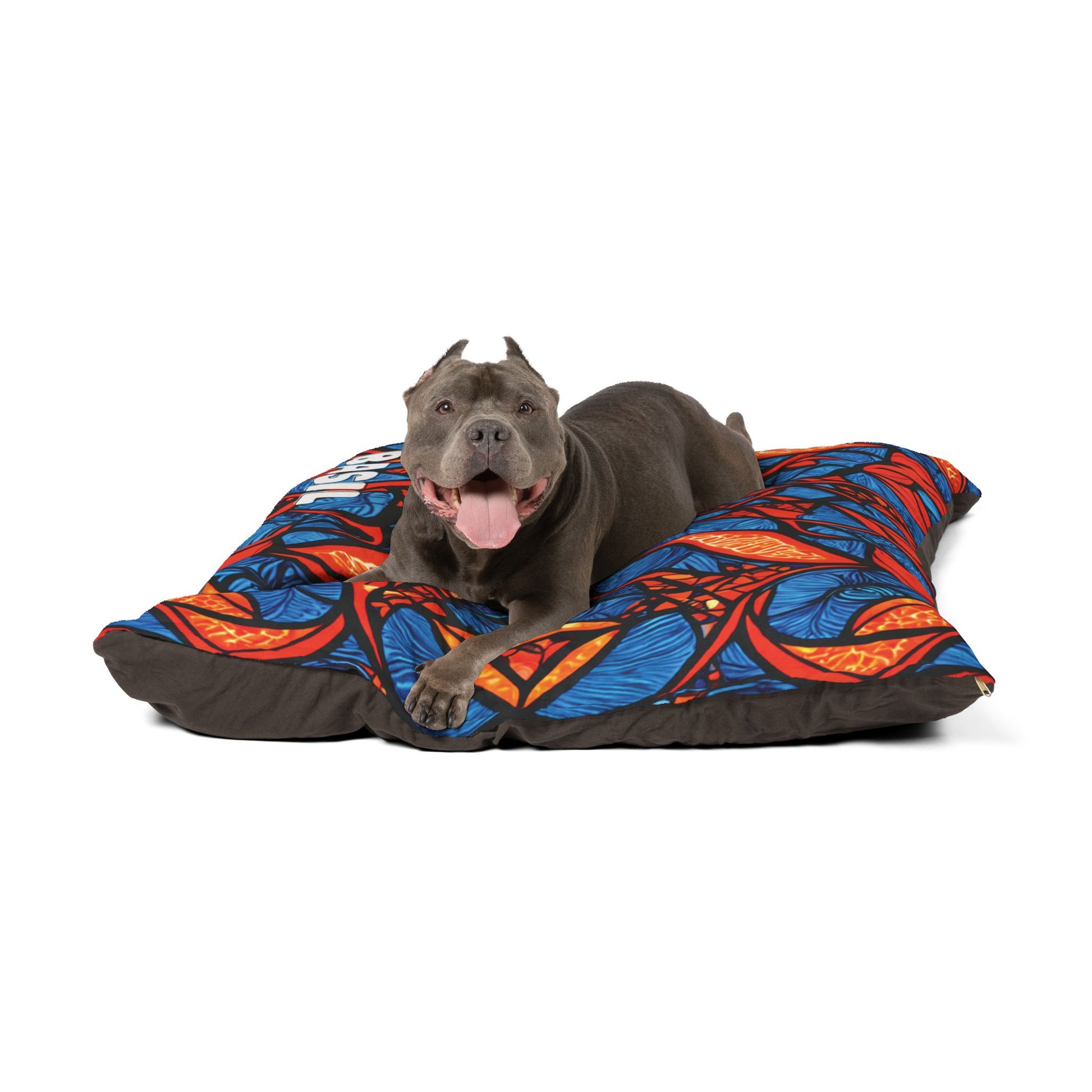 A happy gray pit bull with cropped ears lies on a stylish dog bed featuring a vibrant red, orange, and blue geometric pattern, looking forward with its mouth open and tongue out. The background is plain white.