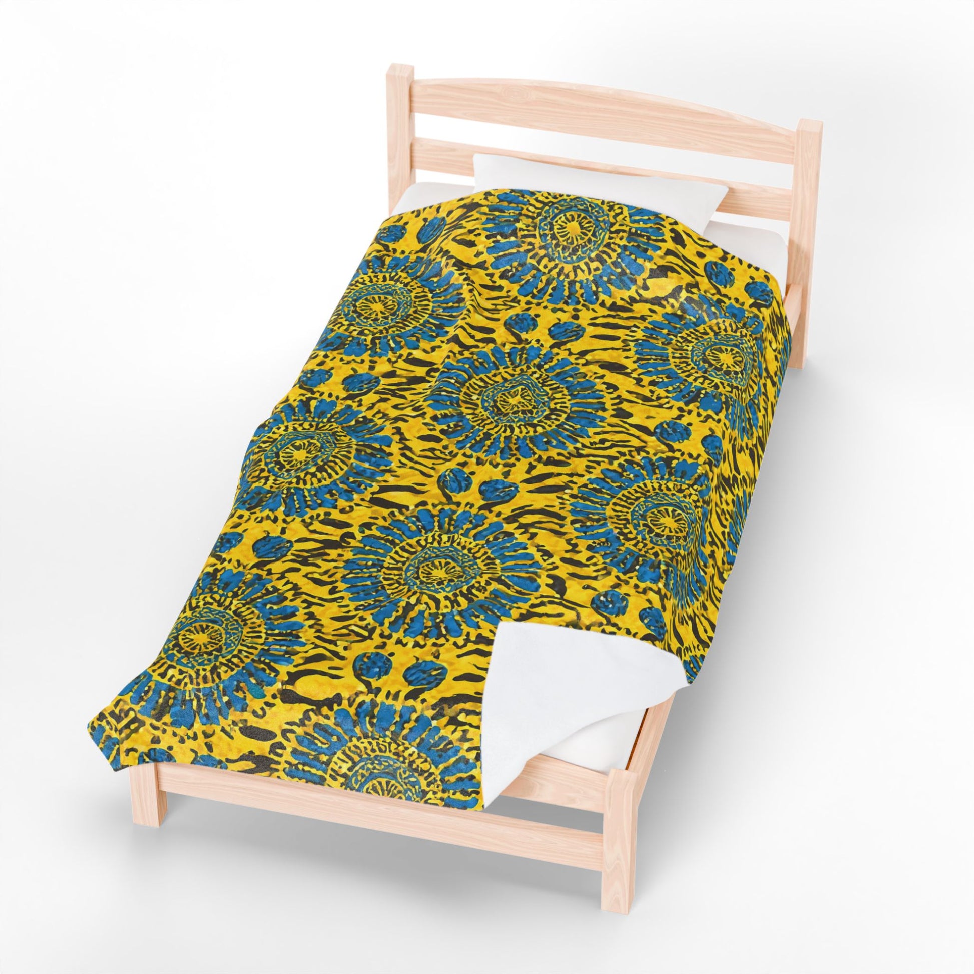 A single wooden bed with white sheets and a vibrant African-inspired blanket featuring a yellow background and bold blue and green circular floral patterns, viewed from above against a plain white backdrop.