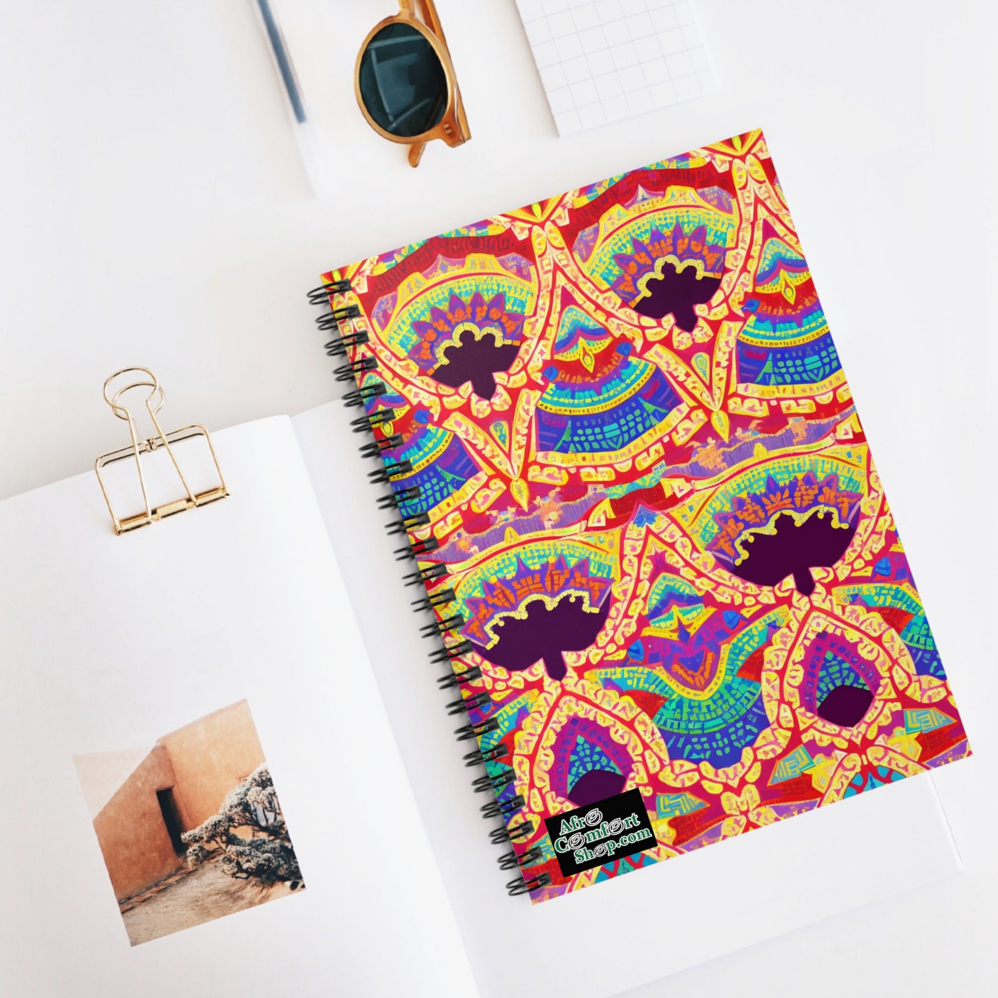 An African-inspired spiral notebook with a colorful, intricate geometric pattern cover rests on an open book displaying a photo of a rustic wall. Nearby are sunglasses, a binder clip, and a small grid notepad, all on a white surface.