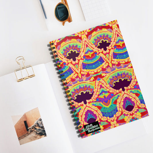 An African-inspired spiral notebook with a colorful, intricate geometric pattern cover rests on an open book displaying a photo of a rustic wall. Nearby are sunglasses, a binder clip, and a small grid notepad, all on a white surface.