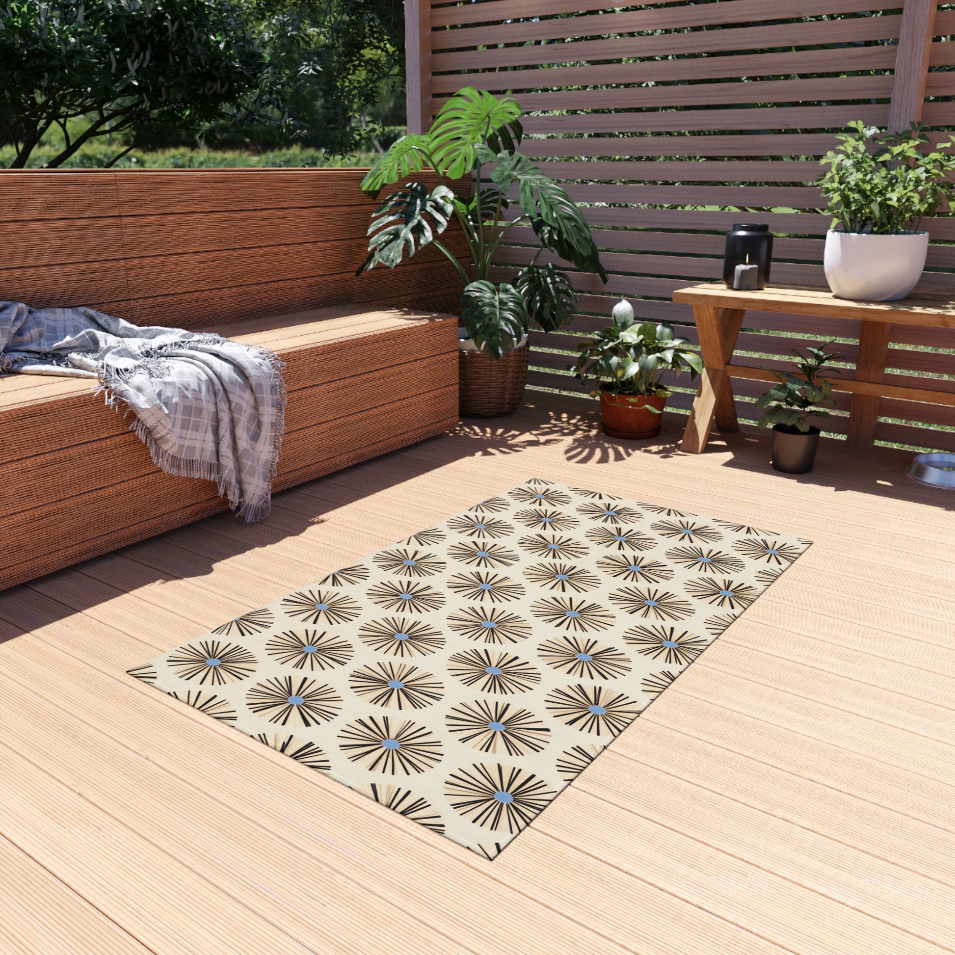A cozy outdoor patio with light wood flooring features an African-inspired outdoor rug in beige and black geometric stars, surrounded by potted plants. A wicker sofa with gray cushions and a plaid throw sits by a wooden privacy screen, bathed in sunlight.
