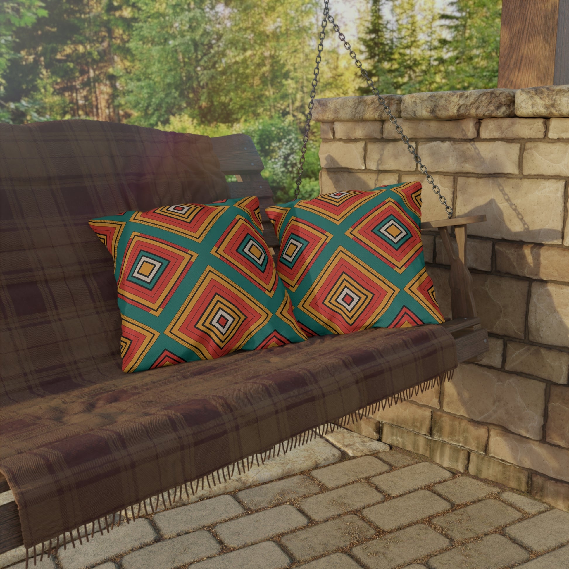 A wooden porch swing with outdoor pillows, a brown plaid blanket, and two colorful diamond-patterned pillows sits by a stone wall. Sunlight filters through nearby trees, casting shadows on the weatherproof materials and stone patio below.