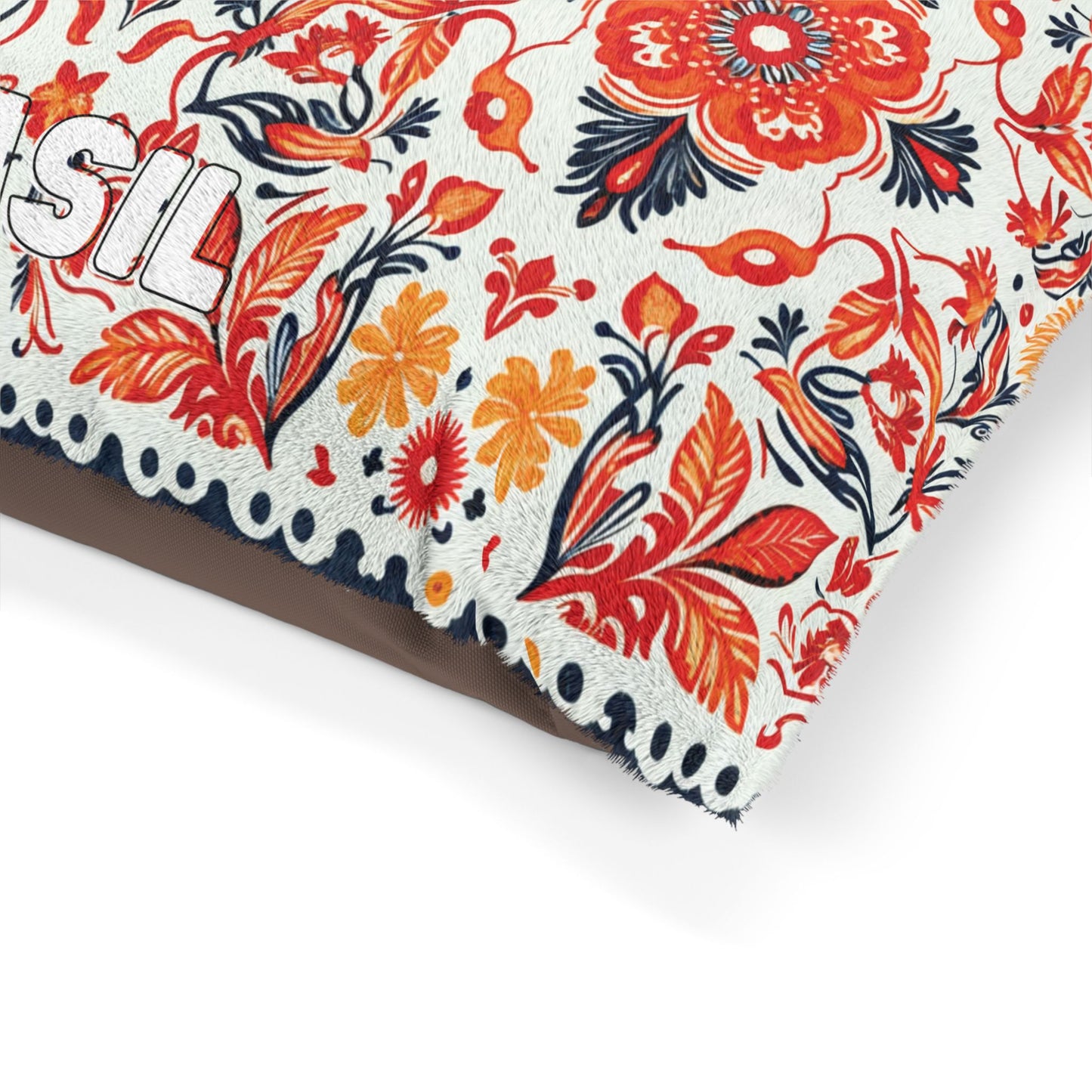 A close-up of a decorative, African-inspired cushion with a cream background, featuring an intricate floral pattern in red, orange, and dark blue. The edge has black stitched dots, and part of the word “SIL” in embroidered white letters is visible.
