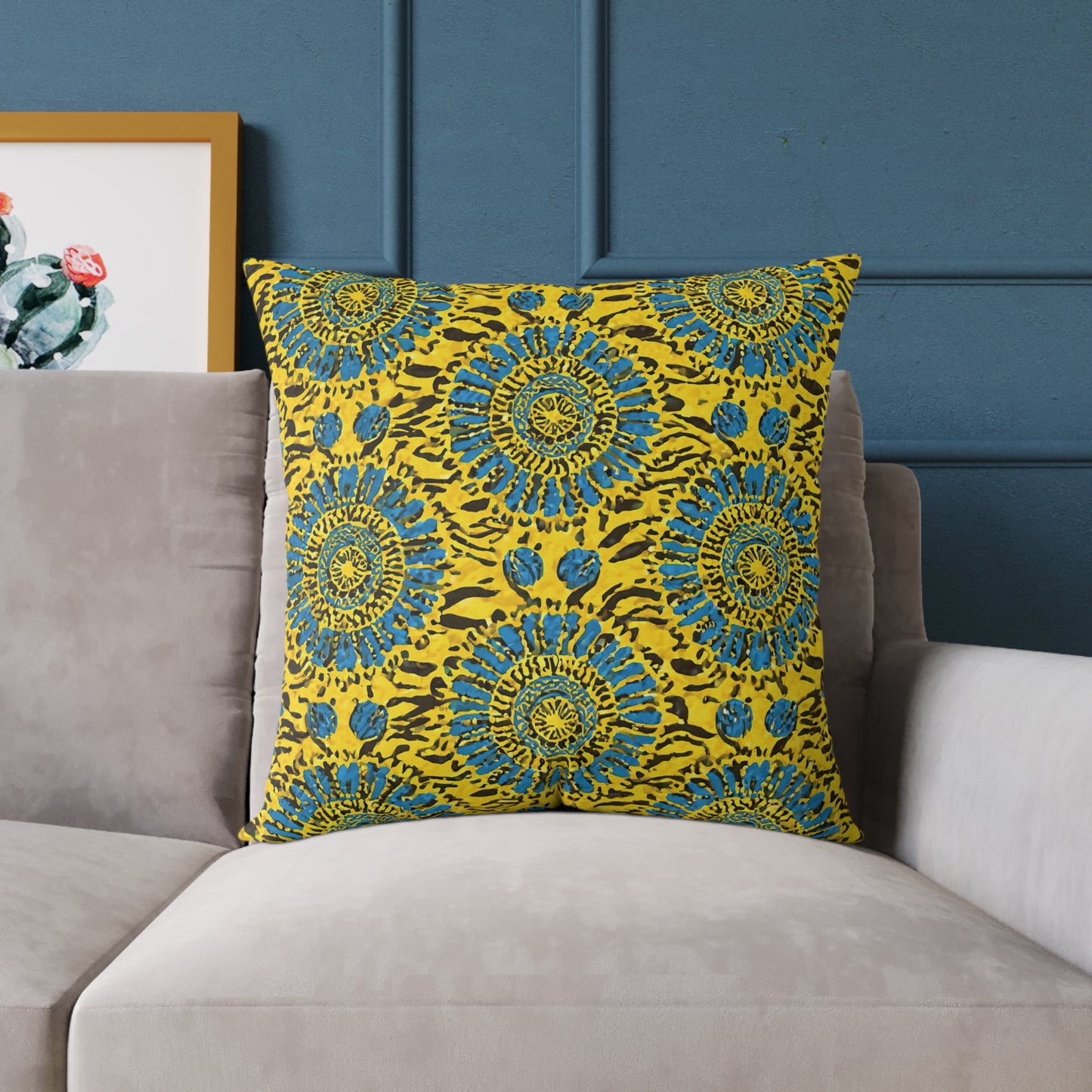 A vibrant accent pillow with a yellow cover features blue and green circular mandala patterns. Placed on a light gray sofa against a blue paneled wall, this African-inspired pillow adds colorful home decor flair beside framed cactus artwork.