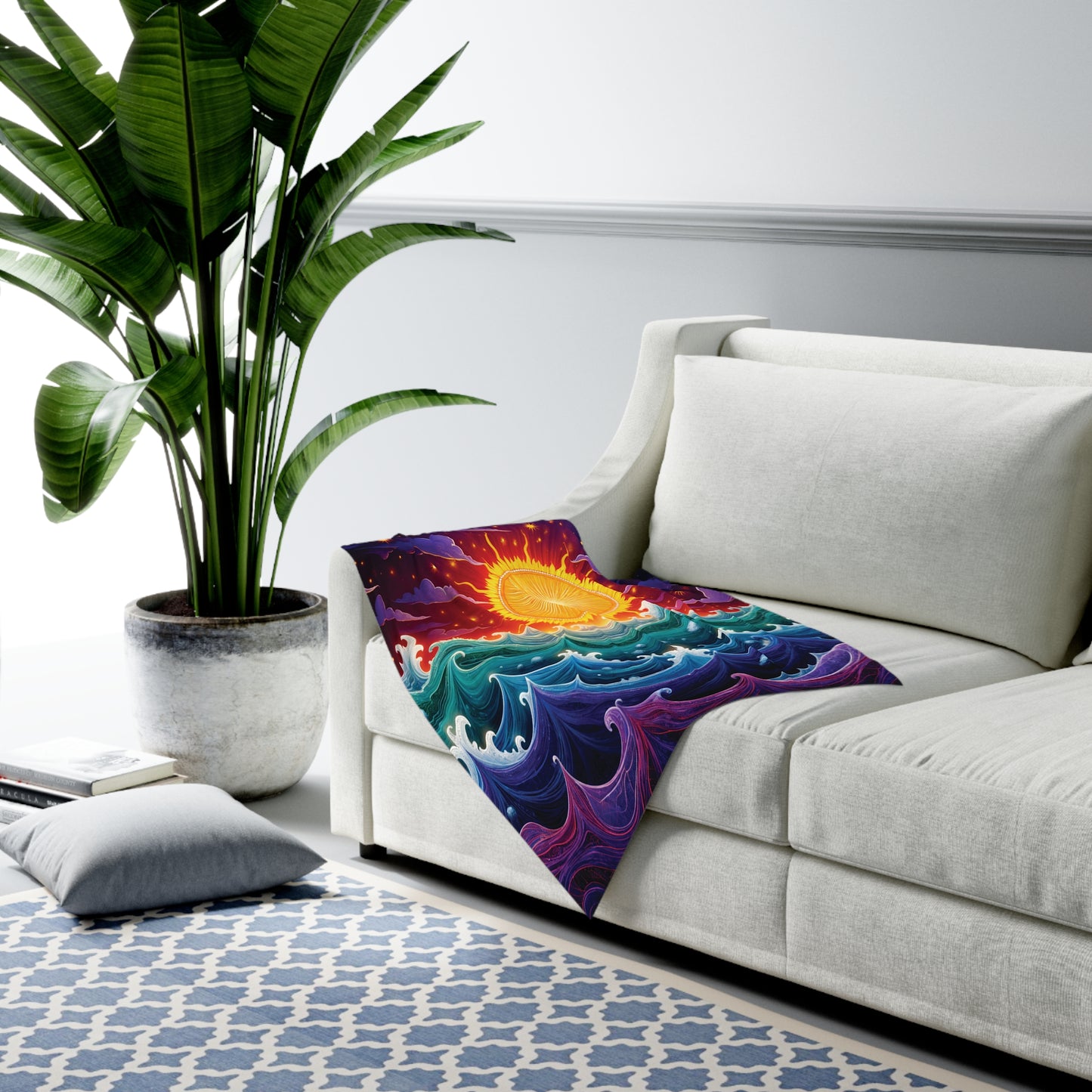 A bright room with a large potted plant, a white sofa, and a colorful customizable baby blanket featuring a vibrant sun over stylized blue and purple waves draped on the couch. A patterned blue rug and a pillow are on the floor nearby.