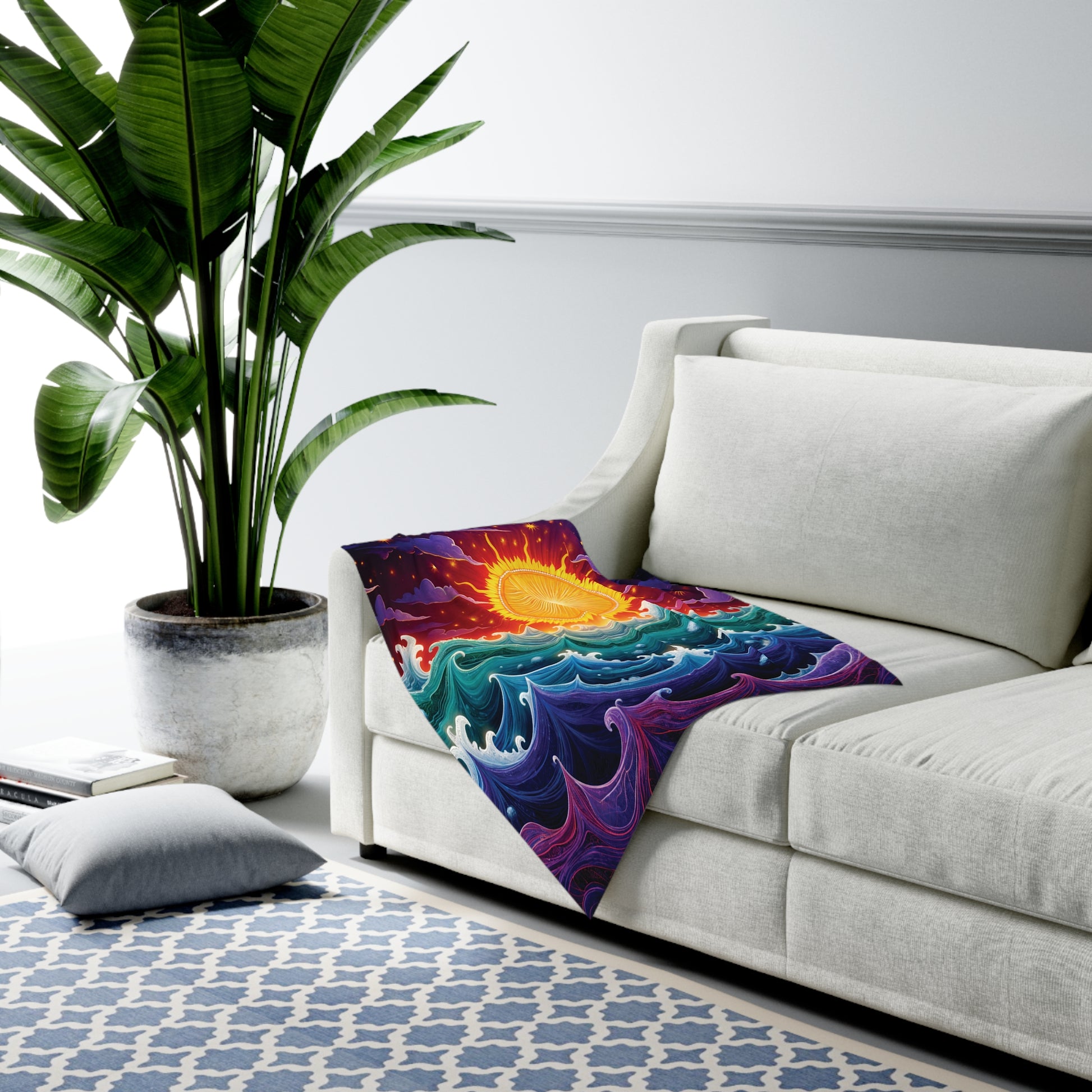 A bright room with a large potted plant, a white sofa, and a colorful customizable baby blanket featuring a vibrant sun over stylized blue and purple waves draped on the couch. A patterned blue rug and a pillow are on the floor nearby.