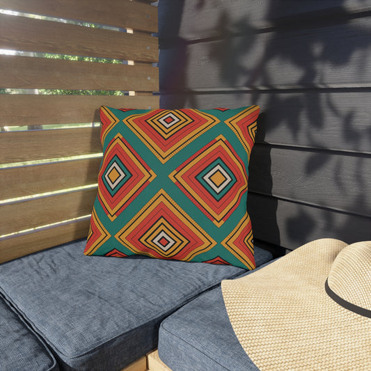 A colorful outdoor pillow, crafted from weatherproof materials with a bold geometric diamond pattern in red, orange, yellow, and teal rests on a blue cushion. Sunlight casts shadows on wooden slats and a straw hat sits nearby, evoking a comfy patio scene.