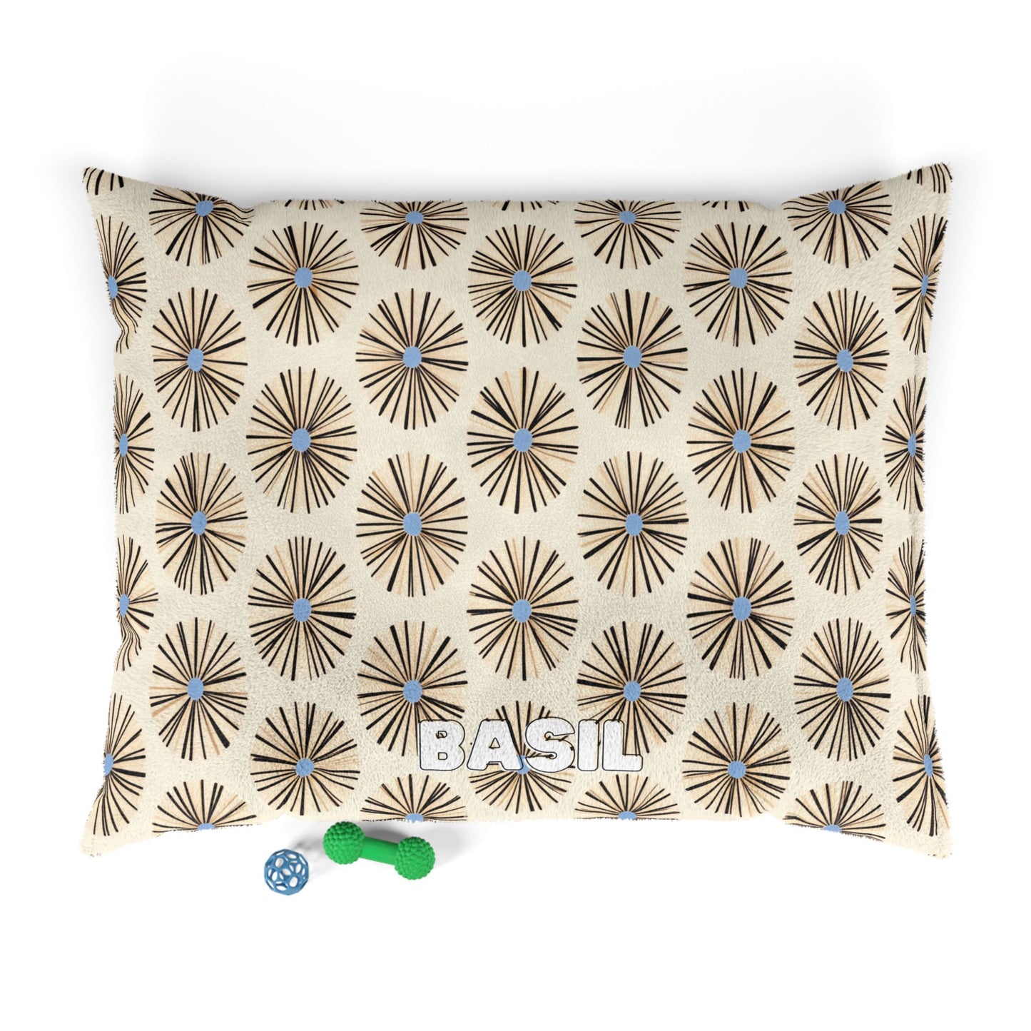 A rectangular pillow with a beige background and an African-inspired brown starburst pattern with blue centers. The name BASIL is printed in bold white letters near the bottom center. Two small pet toys, one green and one blue, sit beside the pillow.