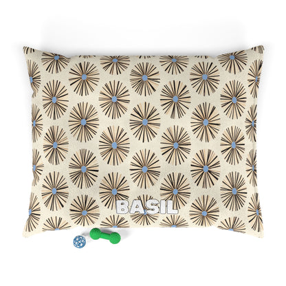 A rectangular pillow with a beige background and an African-inspired brown starburst pattern with blue centers. The name BASIL is printed in bold white letters near the bottom center. Two small pet toys, one green and one blue, sit beside the pillow.