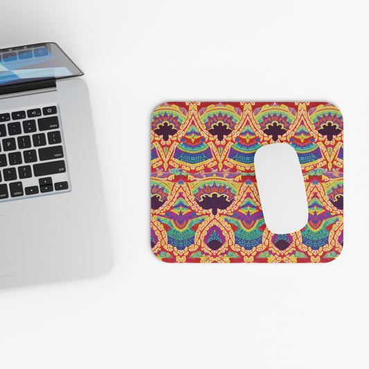 A silver laptop sits on a white surface next to a vibrant mouse pad featuring an intricate, symmetrical geometric pattern in red, orange, blue, and yellow tones. This non-slip desk accessory has a white rectangular area on its right side.