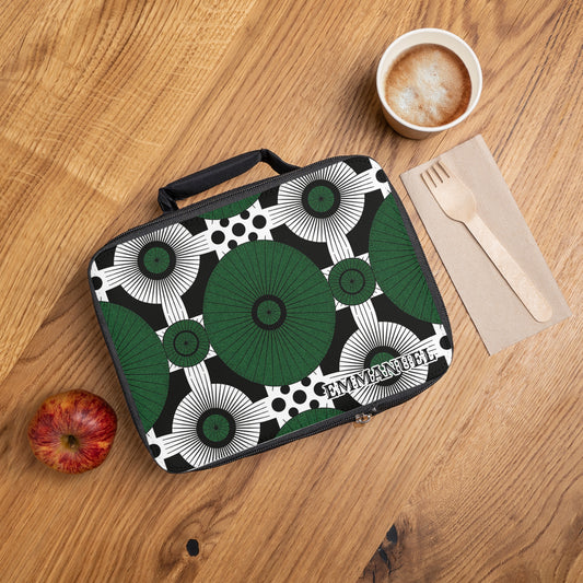 A patterned, African-inspired lunch tote with green and black circular designs and the name EMMANUEL sits on a wooden table next to a red apple, a cup of coffee with foam, and a wooden fork and knife on a brown napkin.