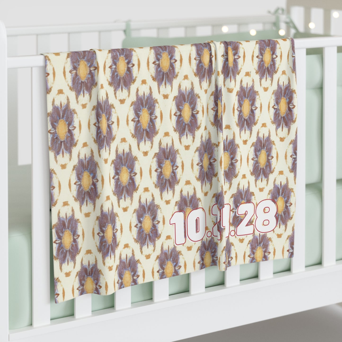 A personalized baby blanket with a repeating pattern of large, purple and yellow flowers hangs over a white crib rail. The date “10.11.28” appears in bold pink and white numbers on the bottom right. The crib mattress is light mint green.