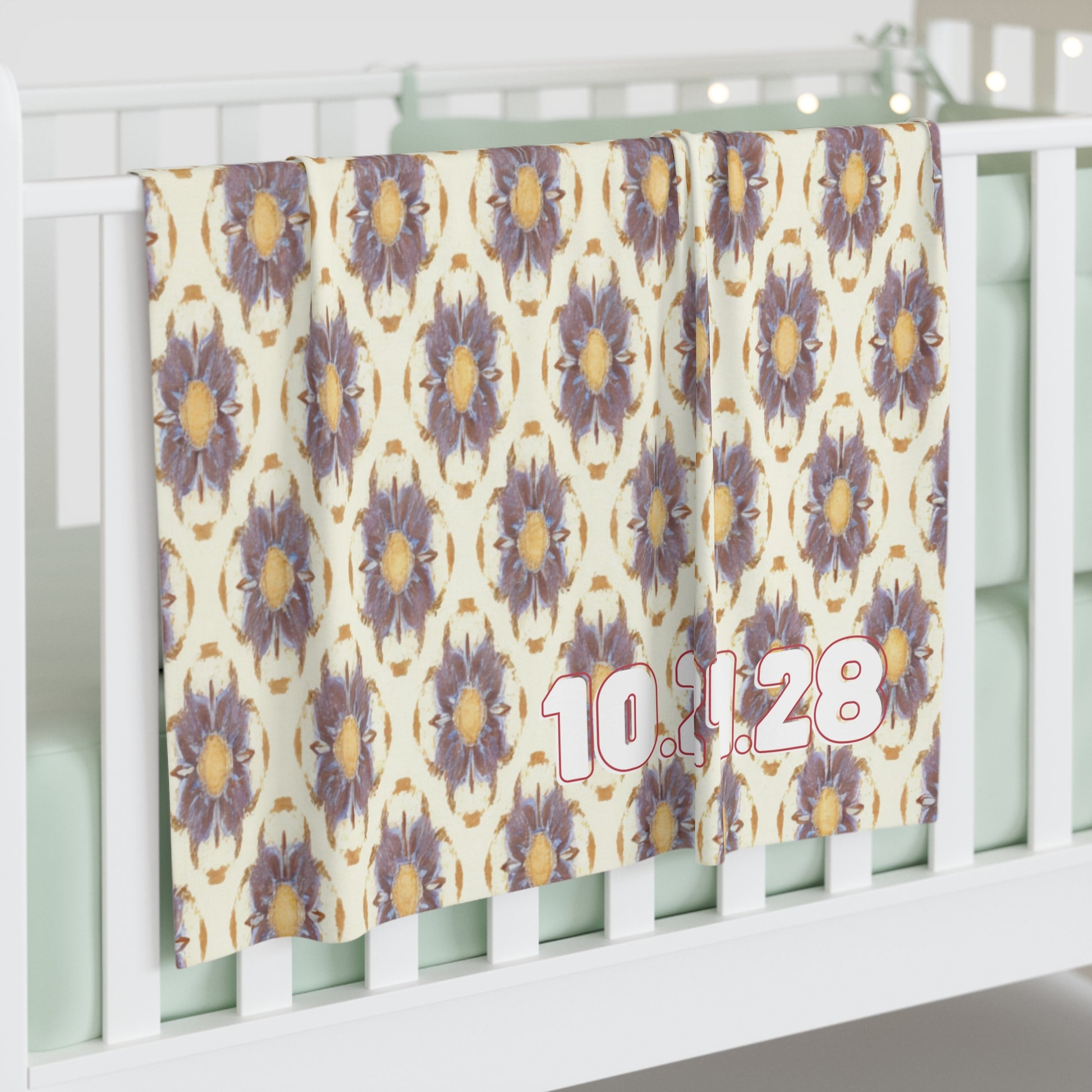 A personalized baby blanket with a repeating pattern of large, purple and yellow flowers hangs over a white crib rail. The date “10.11.28” appears in bold pink and white numbers on the bottom right. The crib mattress is light mint green.