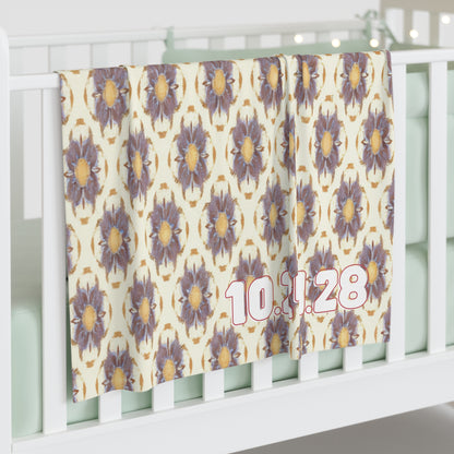 A personalized baby blanket with a repeating pattern of large, purple and yellow flowers hangs over a white crib rail. The date “10.11.28” appears in bold pink and white numbers on the bottom right. The crib mattress is light mint green.