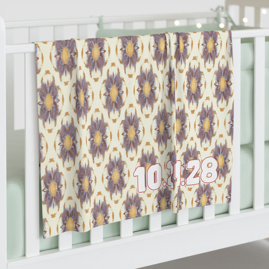 A personalized baby blanket with a repeating pattern of large, purple and yellow flowers hangs over a white crib rail. The date “10.11.28” appears in bold pink and white numbers on the bottom right. The crib mattress is light mint green.