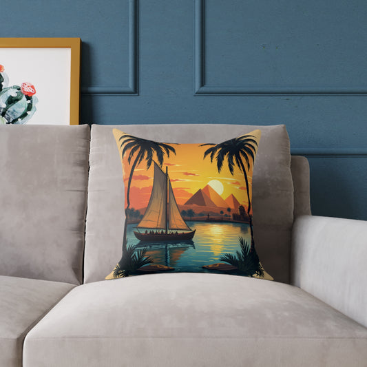 A beige sofa with a colorful accent pillow featuring a sunset scene: a sailboat on calm water, palm trees, distant pyramids, and an orange sky. A framed cactus print is partially visible on the blue wall behind the sofa.