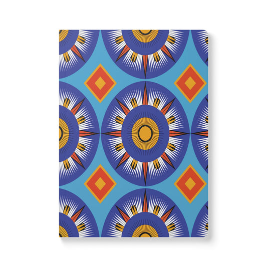 A vibrant African-inspired journal with a geometric cover of blue circular sunburst patterns and bold red and orange diamonds on a blue background—perfect for daily journaling. The notebook’s edge casts a subtle shadow.