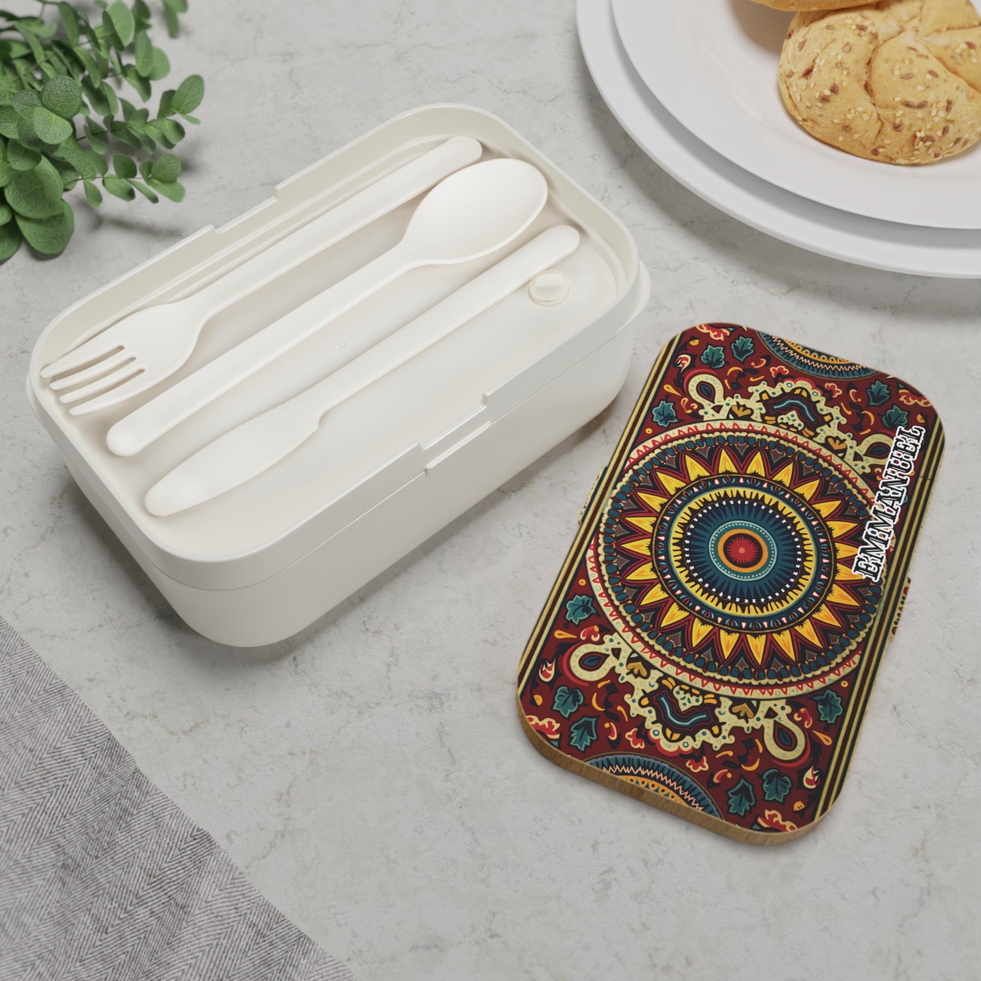 A white BPA-free lunchbox with a fork, spoon, and knife sits on a marble surface next to a vibrantly patterned rectangular tin labeled “IMANIGINE.” A plate of cookies, a green plant, and a gray napkin are nearby.