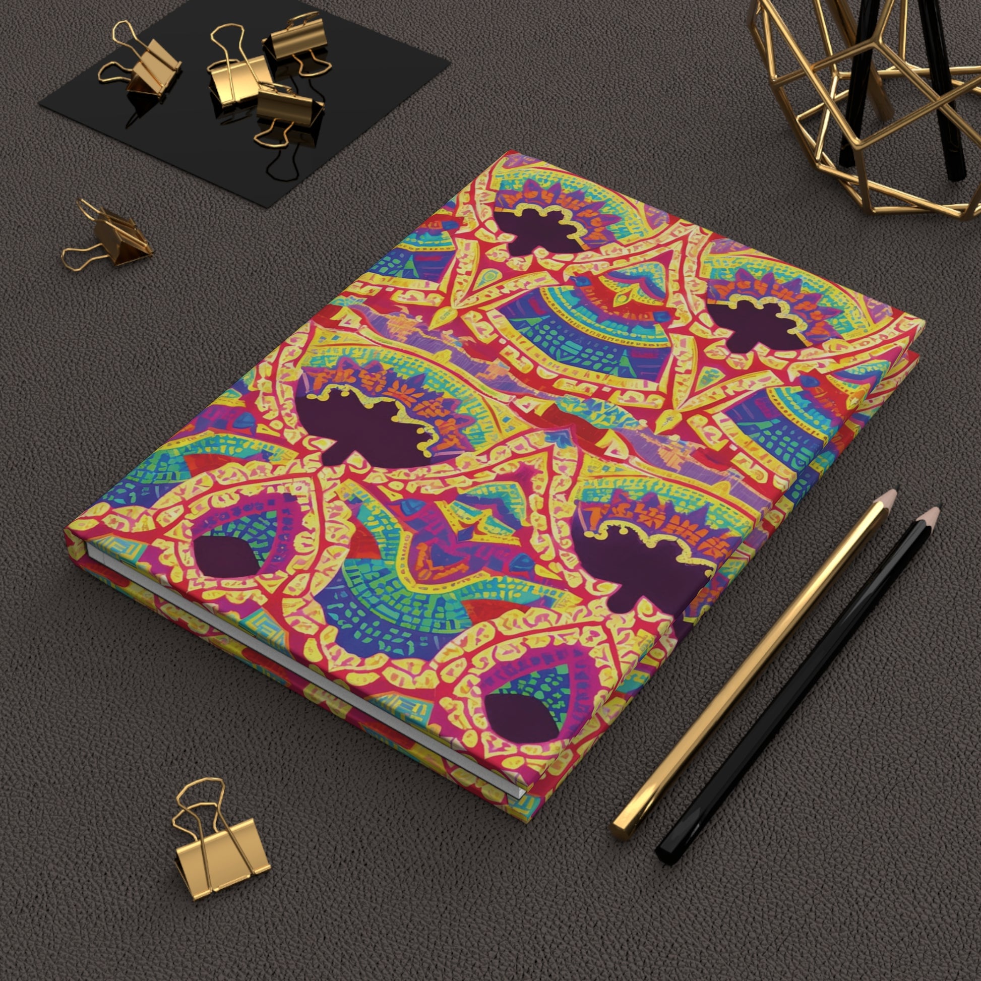 A colorful hardcover journal with intricate, African-inspired geometric and abstract patterns in red, yellow, green, blue, and purple lies on a textured dark brown surface. Nearby are a black and gold pen, pencil, binder clips, and a geometric desk accessory.