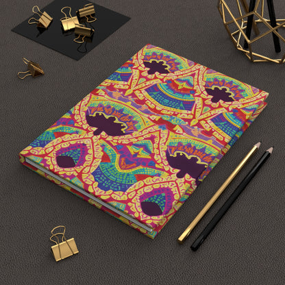 A colorful hardcover journal with intricate, African-inspired geometric and abstract patterns in red, yellow, green, blue, and purple lies on a textured dark brown surface. Nearby are a black and gold pen, pencil, binder clips, and a geometric desk accessory.