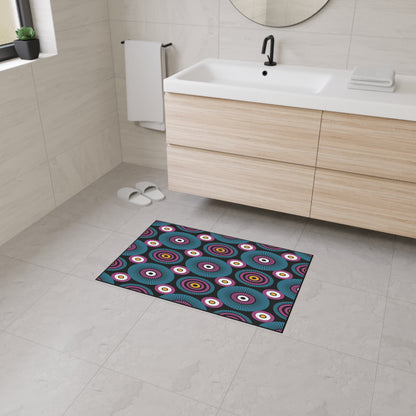 A modern bathroom with light beige tiles, a floating wood vanity, white sink, round mirror, and black faucet. The floor features an African-inspired non-slip rug with concentric circles and geometric patterns in bold colors.
