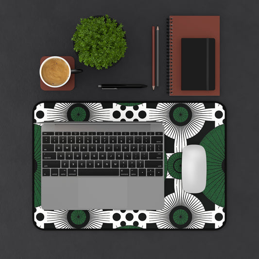 A laptop and white mouse rest on an ergonomic desk mat with black, white, and green patterns. Beside them sit a brown notebook, pencil, black pen, small potted plant, and a coffee cup—all arranged on a dark surface.