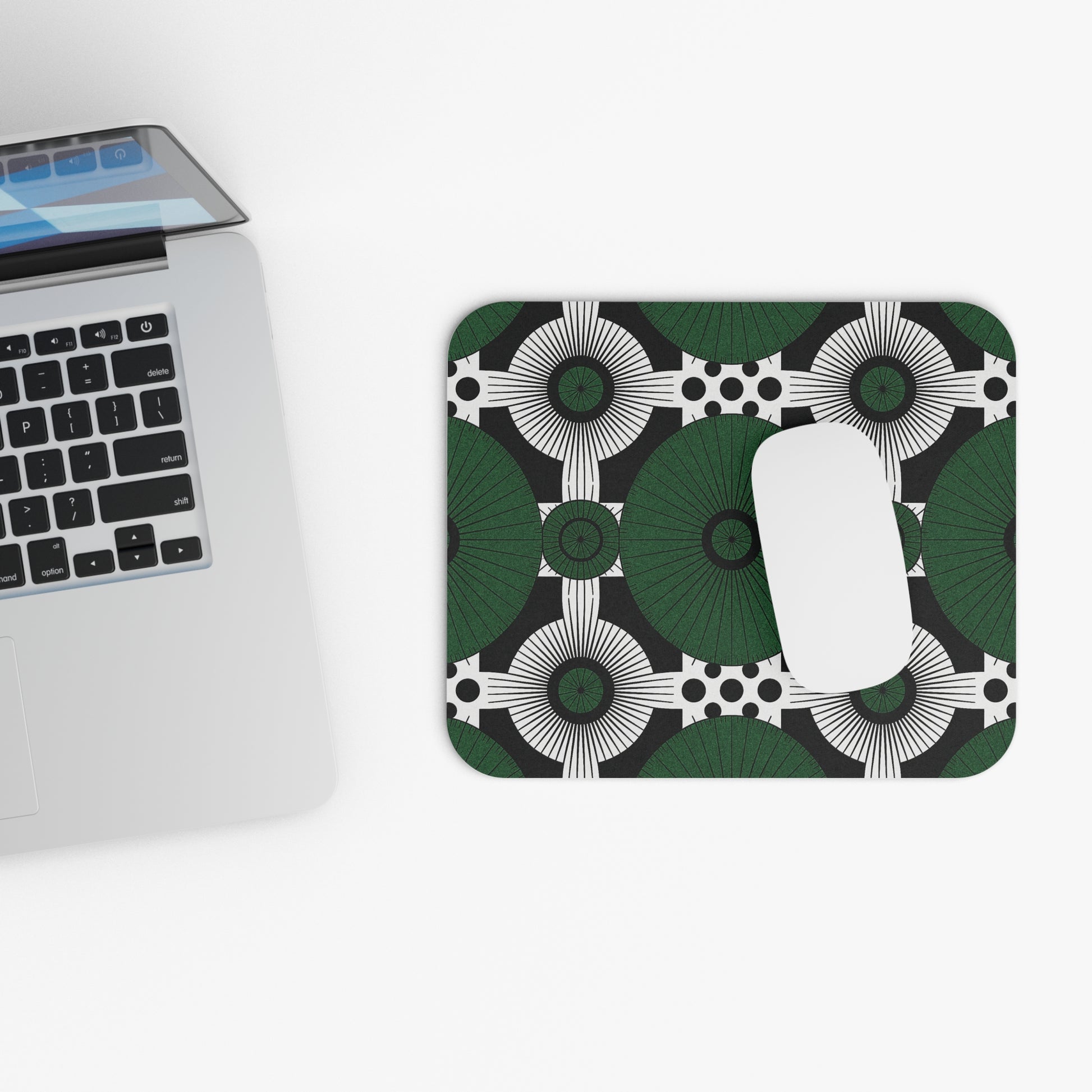 A silver laptop sits on a white surface next to an African-inspired mouse pad featuring green, black, and white circles. This non-slip desk accessory boasts durable high-density foam, with a white wireless mouse resting on top.