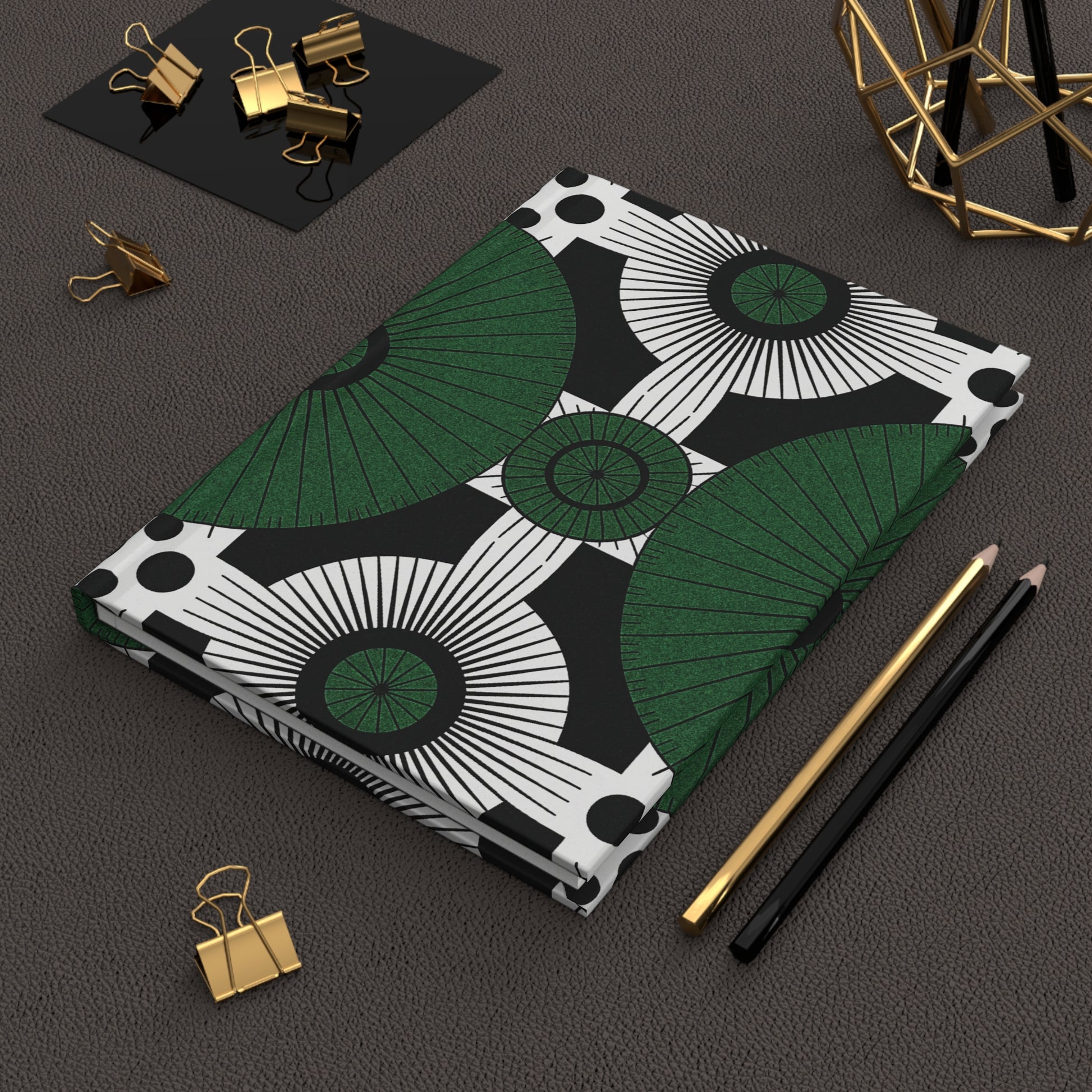 A hardcover notebook with a bold geometric green, black, and white pattern lies on a textured brown surface. Beside it are a gold pen, a black pencil, gold binder clips, and a decorative wire ornament for a modern workspace.